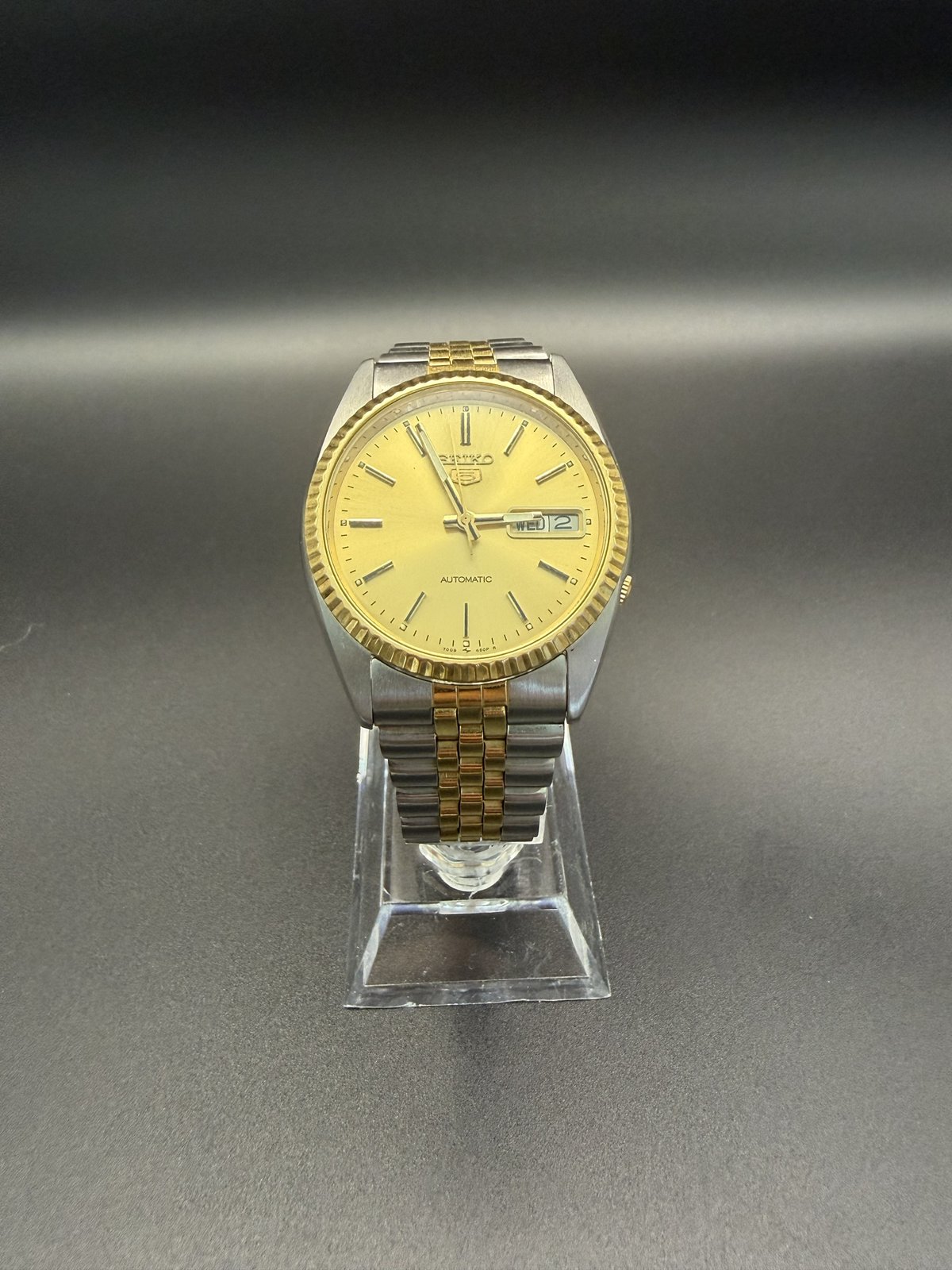 Seiko 5 automatic SNXJ92 with gold dial and two-tone bracelet, front view on a stand